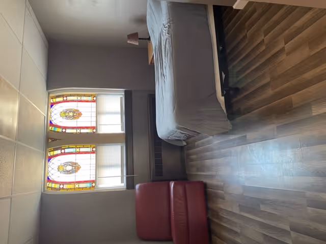 A simple bedroom with a single bed covered in a gray bedspread, a small wooden nightstand with a lamp, a red armchair, and two stained glass windows with colorful designs. The room has wood-patterned flooring and gray walls.