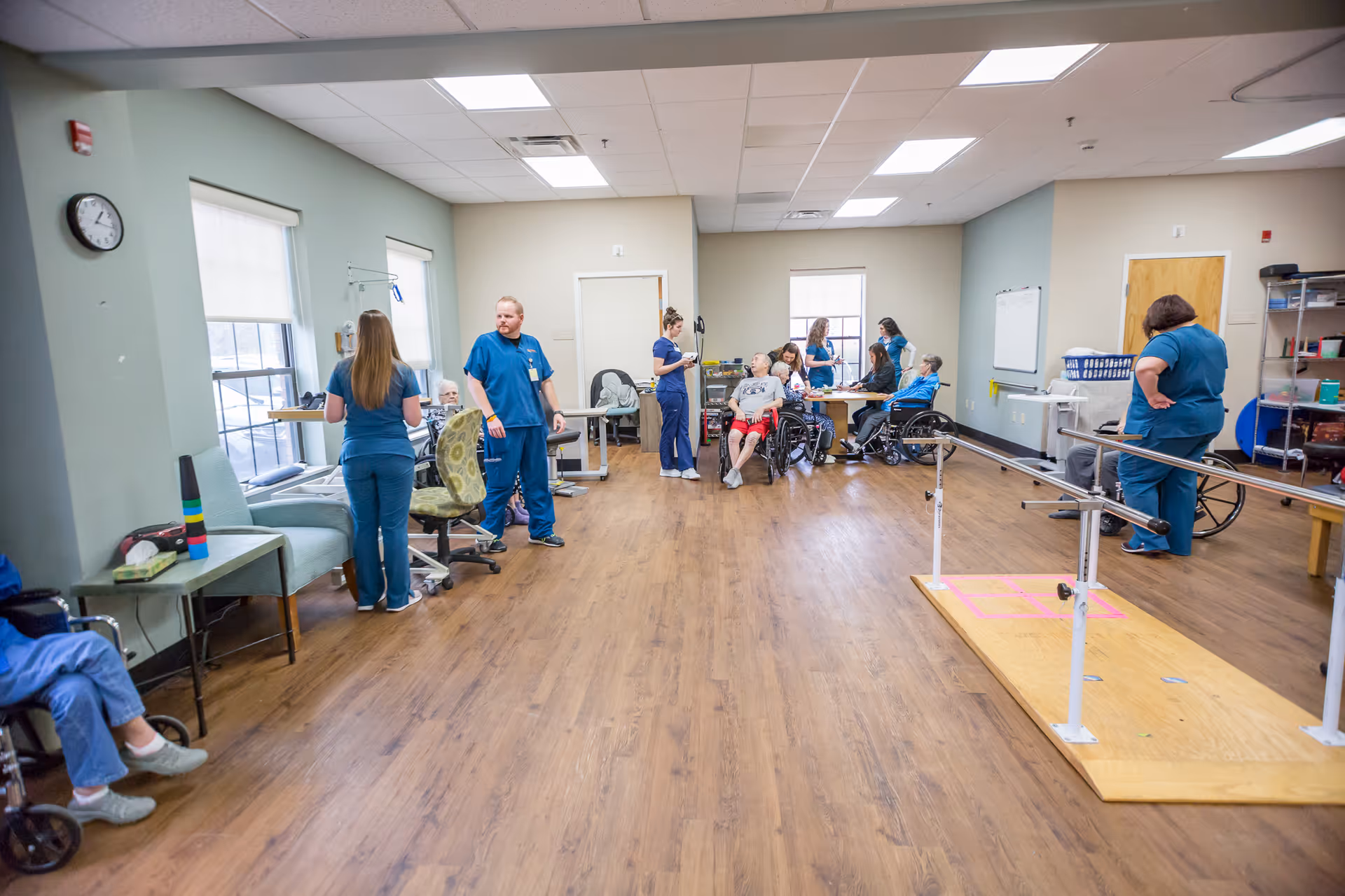 A spacious room in a senior care facility with several elderly individuals, some in wheelchairs, accompanied by healthcare staff in blue scrubs. The room has wooden flooring, large windows with blinds, and various medical and therapy equipment including parallel bars for walking exercises. The atmosphere appears to be a therapy or rehabilitation session.