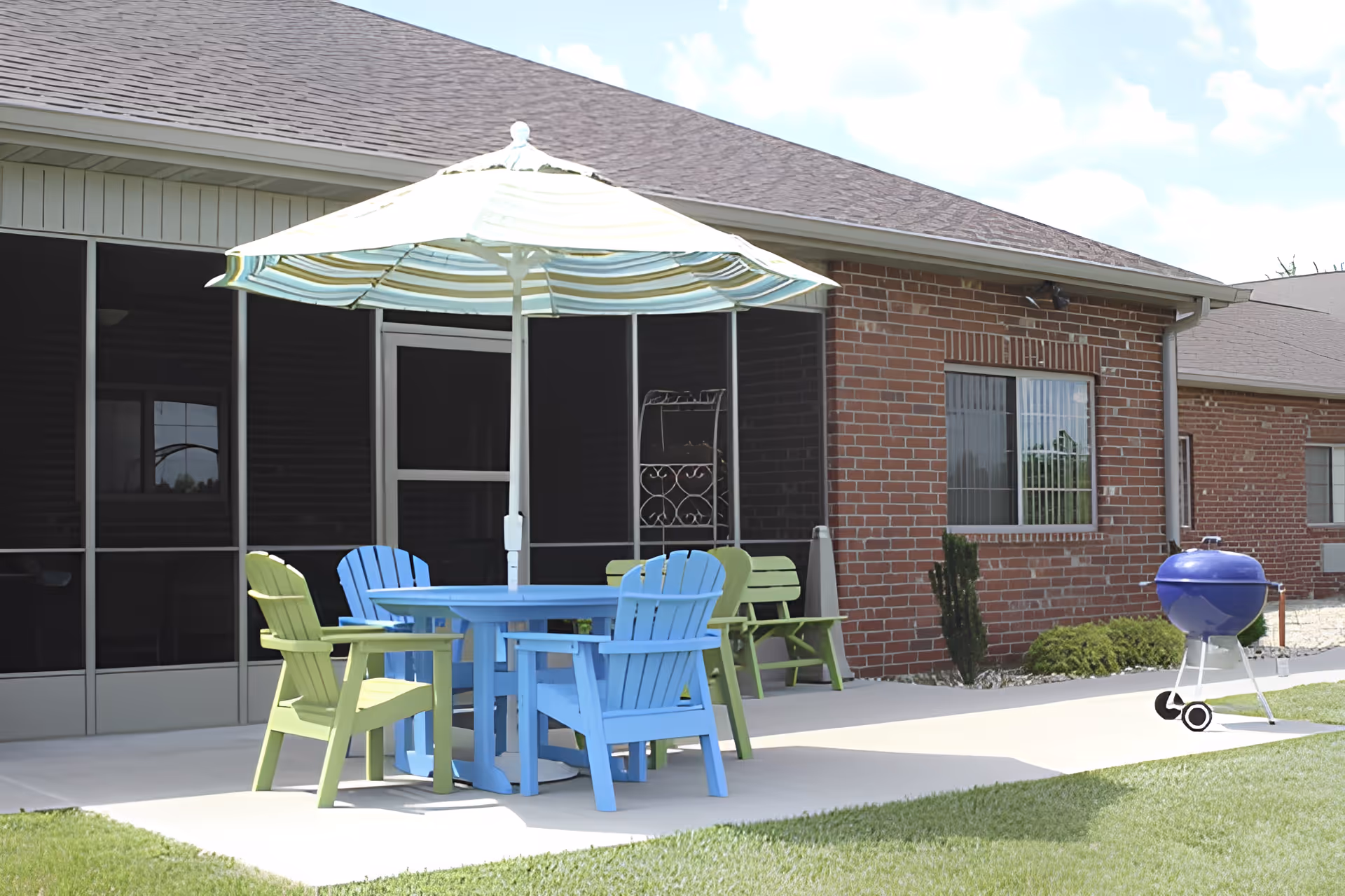 Patio area with a striped umbrella over a blue table and colorful chairs on a concrete slab beside a brick building.