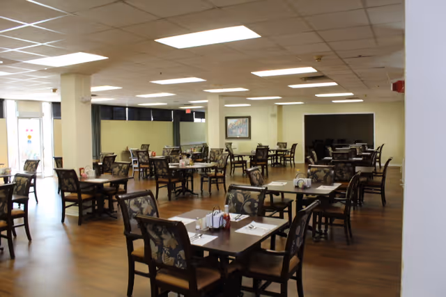 A spacious dining room with multiple dark wood tables and chairs arranged neatly. Each table is set with placemats, napkins, and condiments. The room has a wooden floor, light-colored walls, and a drop ceiling with fluorescent lighting. There is a framed picture on one wall and large windows with black blinds on another side.