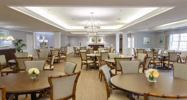 Spacious communal dining room with multiple round wooden tables and upholstered chairs, chandeliers, and small floral centerpieces.