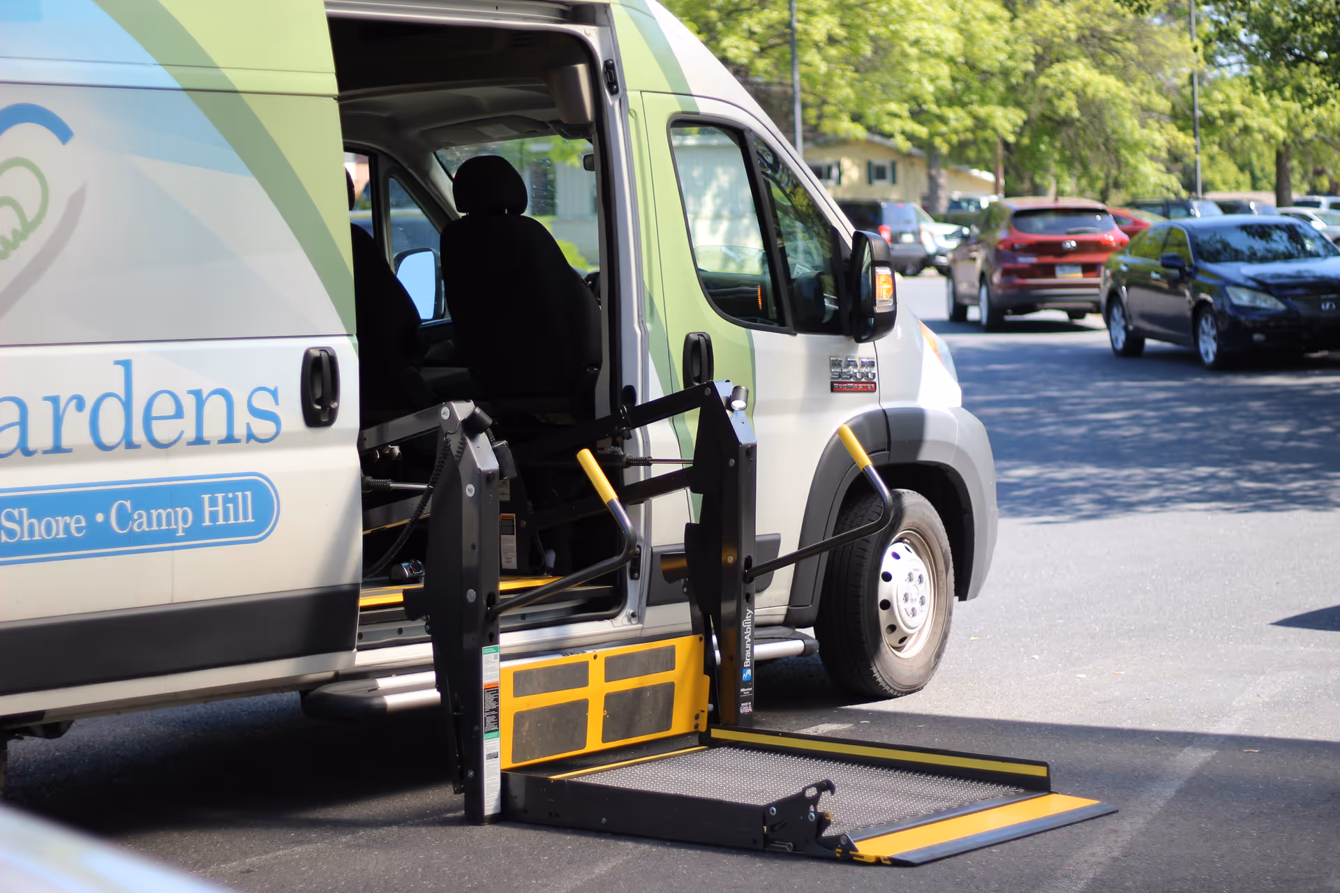 A white and green van with an open side door showing a wheelchair lift extended to the ground in a parking lot with several cars and trees in the background.