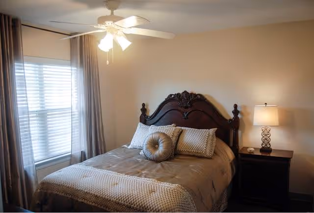 A cozy bedroom with a large wooden headboard bed, decorated with multiple pillows and a beige comforter. There is a nightstand with a decorative lamp on the right side of the bed, and a window with blinds and curtains on the left side. A ceiling fan with lights is mounted on the ceiling.