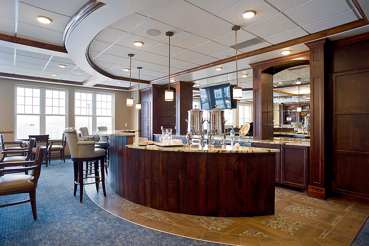 Interior view of a senior living facility's common area featuring a curved wooden bar with beverage dispensers, high chairs, pendant lights, large windows letting in natural light, and a carpeted floor with tile accents.