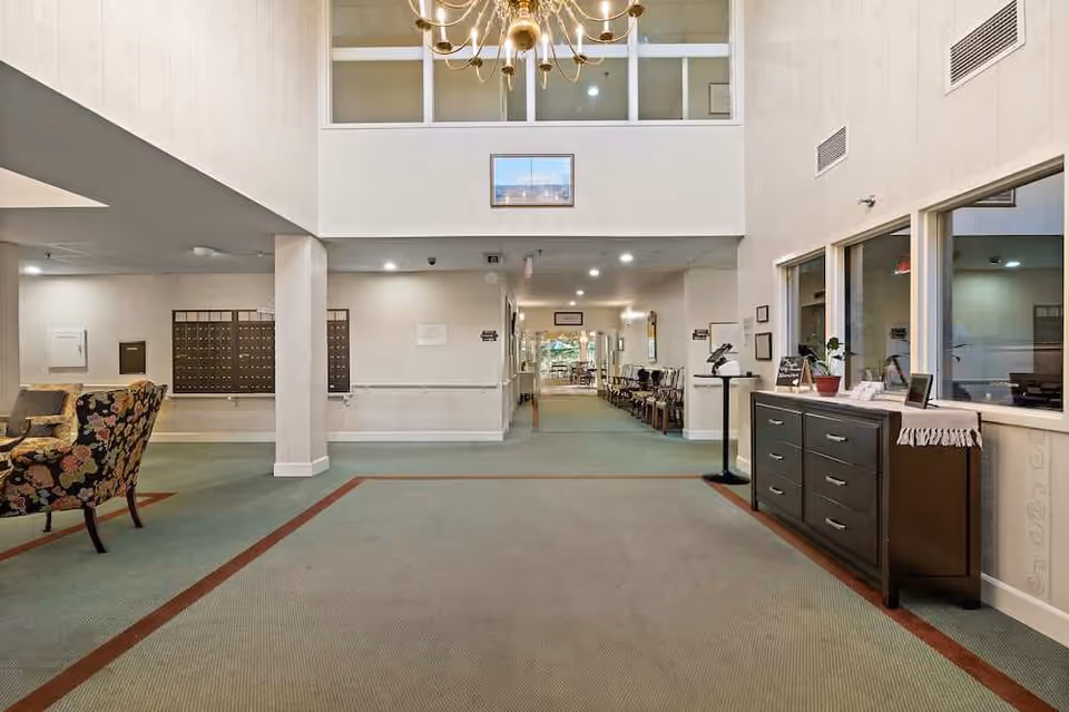 Bright two-story senior living lobby with seating, mailboxes, a chandelier, and a hallway leading to communal spaces.