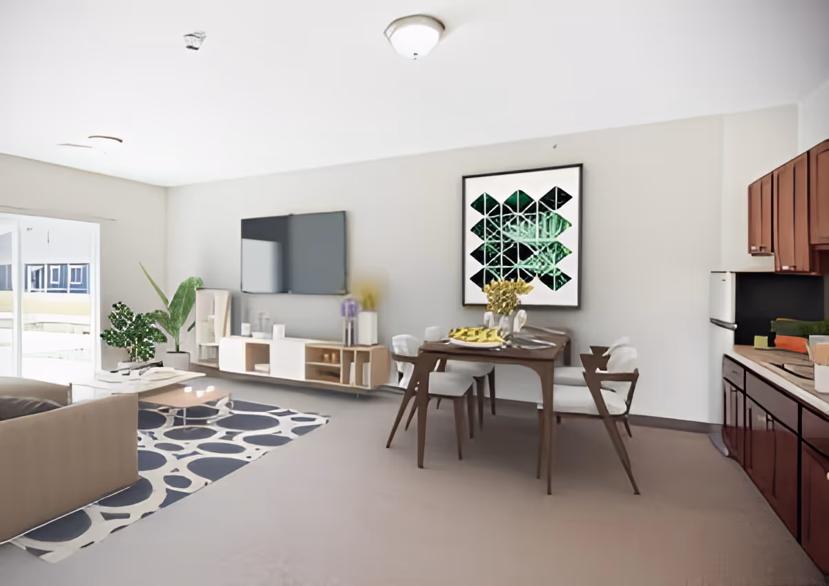 A bright and modern living and dining area in an assisted living facility. The room features a beige sofa, a patterned rug, a wooden coffee table, a wall-mounted flat-screen TV above a low white and wood media console, and a dining table with four chairs. There is a large framed geometric artwork on the wall and a kitchen area with wooden cabinets and a refrigerator on the right side. Large windows allow natural light to fill the space.