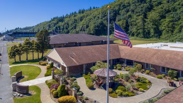 Aerial view of Pacific Care Center showing a single-story building with a brown roof surrounding a landscaped courtyard with a gazebo and various shrubs. An American flag on a flagpole is prominently displayed in the courtyard. The facility is set against a backdrop of green wooded hills under a clear blue sky.