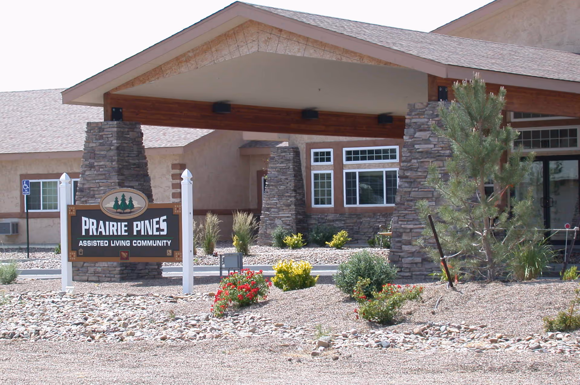 Entrance of Prairie Pines Assisted Living Community featuring a covered driveway supported by stone pillars, a sign with the facility name, and landscaped area with shrubs and flowers.