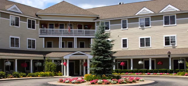 Front exterior of a three-story beige senior living building with balconies, a covered main entrance, circular driveway and landscaped flower beds.