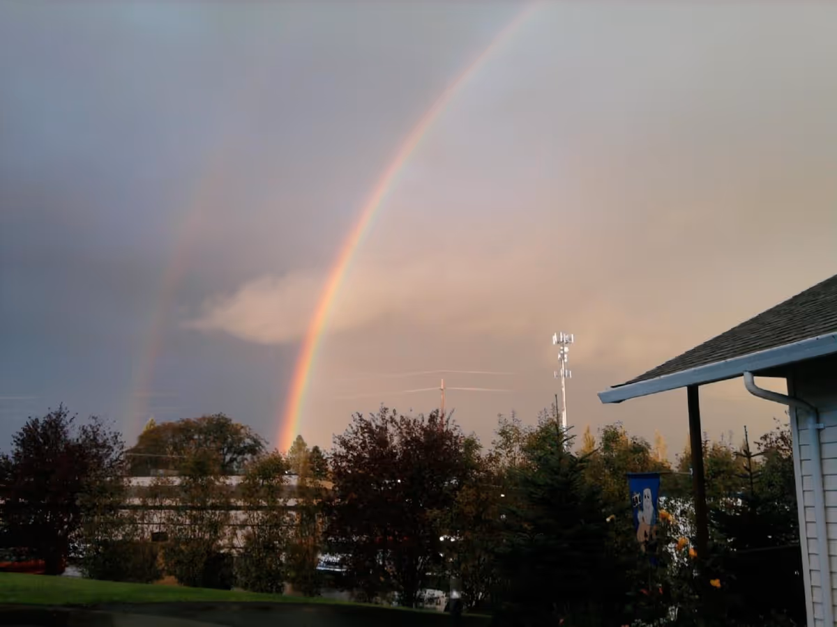A double rainbow appears in the sky above a landscape with trees, a grassy area, and part of a building with a roof and gutter visible on the right side.
