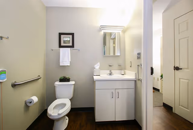 A clean and simple bathroom with a white toilet, a white vanity with a sink, a mirror above the sink, a towel rack with a white towel, a small framed picture on the wall, and a soap dispenser mounted on the wall. The bathroom has light-colored walls and dark wood flooring. The door to an adjacent room is open, showing part of a carpeted area.
