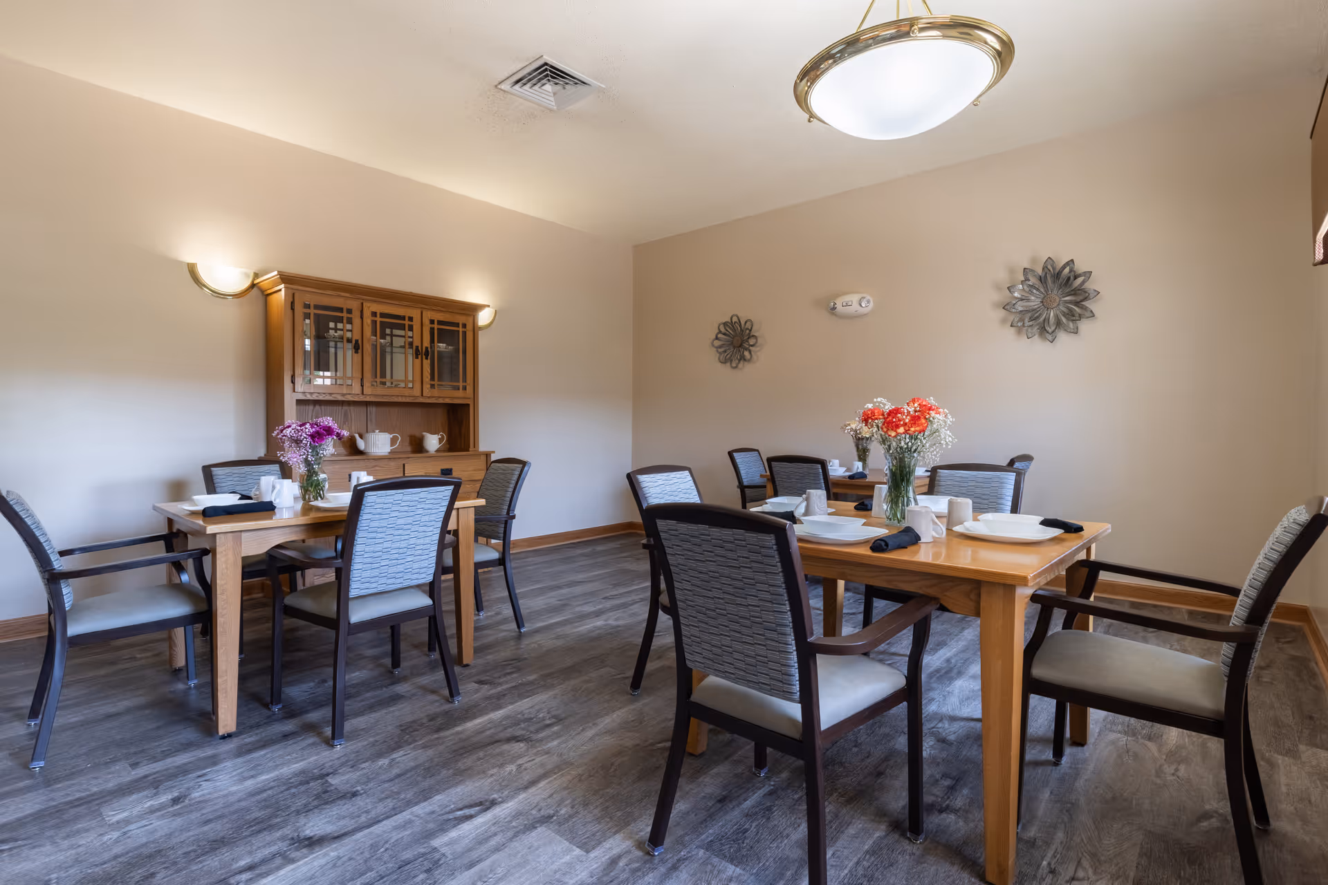 Dining room with wooden tables set for meals, upholstered chairs, floral centerpieces, and a wooden cabinet against the wall.