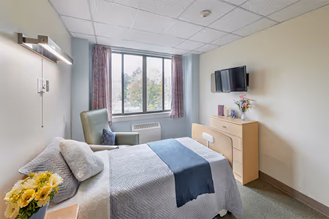 A bright and clean bedroom in a senior living facility with a neatly made bed featuring gray and blue bedding, a green armchair with a pillow, a wooden dresser with flowers and books, a wall-mounted flat-screen TV, and a large window with patterned curtains letting in natural light.