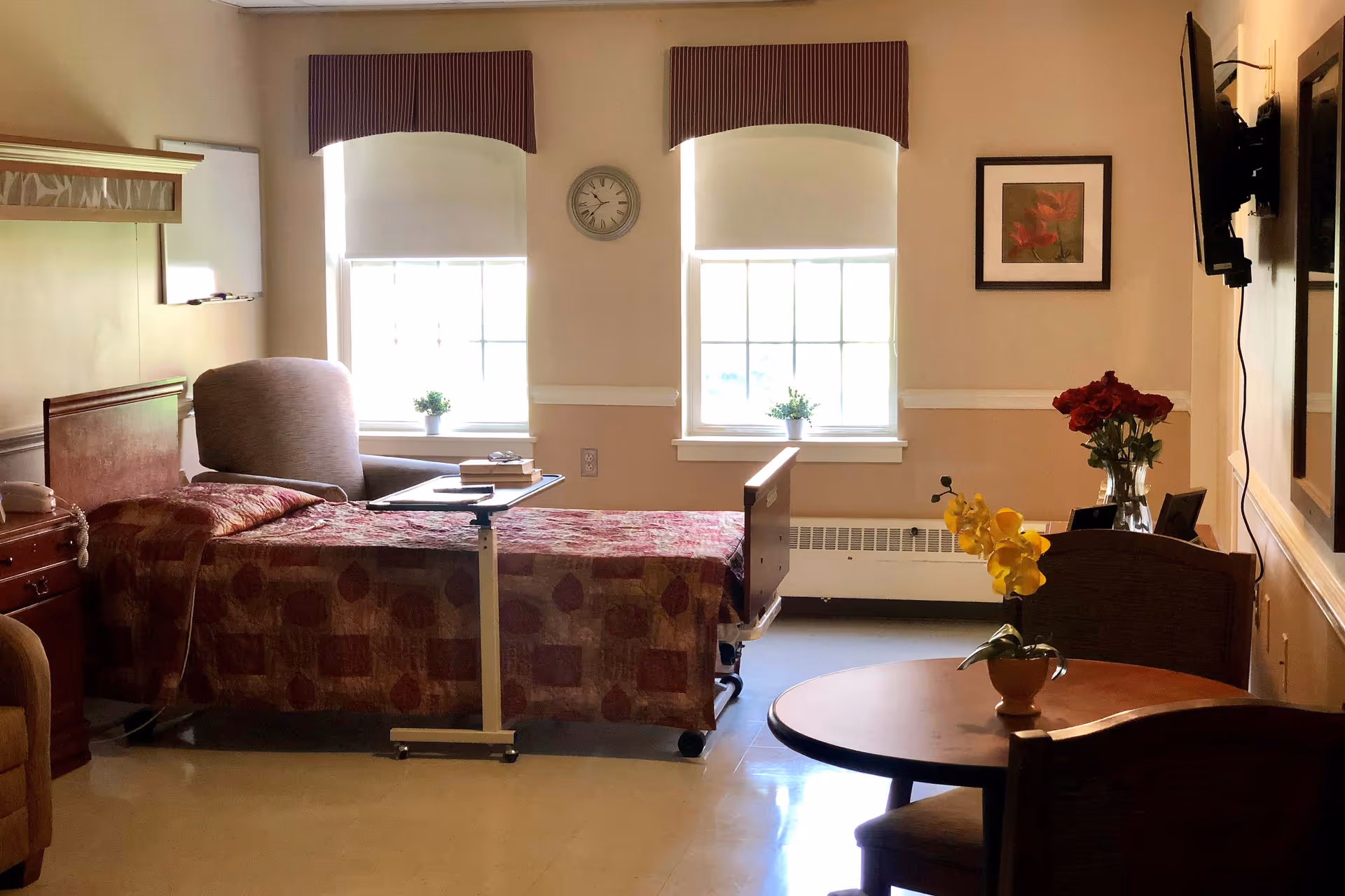 A well-lit bedroom in a senior living facility with a single bed covered in a patterned quilt, a comfortable armchair, a small overbed table with books, two windows with blinds and valances, a wall clock, a framed floral picture, a wall-mounted TV, and a round table with chairs and flower vases.