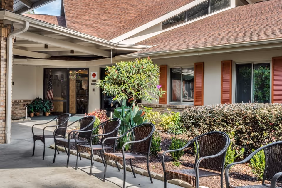 Outdoor seating area at the entrance of a senior living facility with several empty wicker chairs arranged along a covered walkway. There are plants and shrubs along the building wall with windows and red shutters. The entrance door is glass with an American flag visible inside.