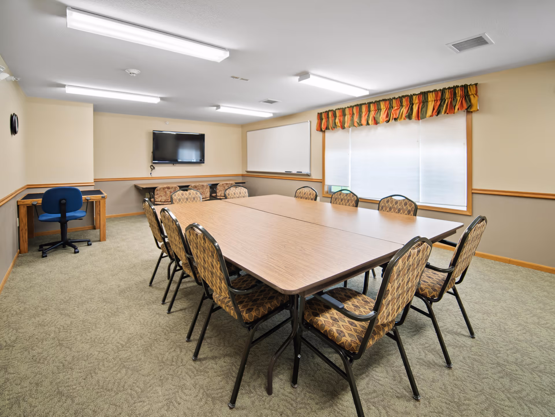 A meeting or conference room with a large rectangular table surrounded by ten patterned chairs. The room has beige walls with a chair rail, a carpeted floor, a wall-mounted flat screen TV, a whiteboard, and a window with blinds and a colorful valance. There is also a small desk with a blue office chair in the corner.
