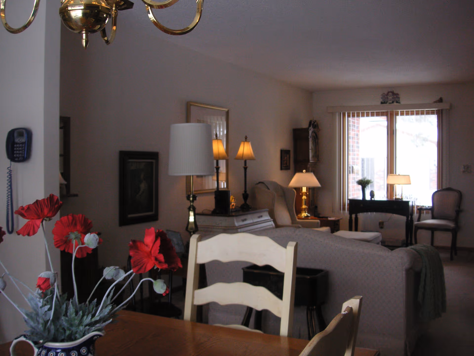 Interior view of a senior living facility's common area featuring a dining table with a vase of red flowers in the foreground, a beige couch, armchairs, multiple lamps, framed artwork on the walls, and large windows with vertical blinds letting in natural light.