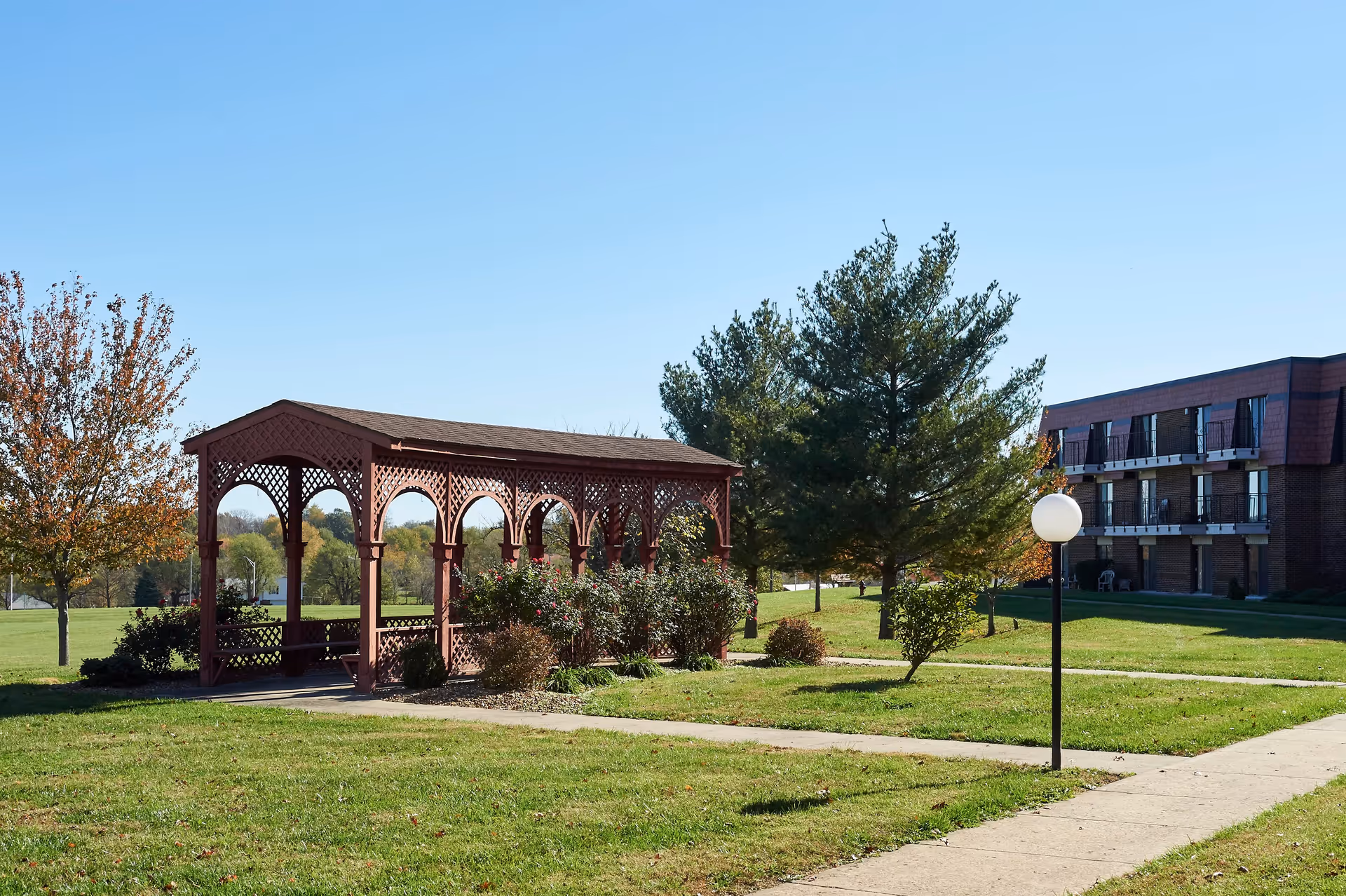 A red wooden gazebo with lattice design stands on a grassy lawn surrounded by bushes and trees with autumn leaves. A paved walkway leads to the gazebo and a three-story brick building with balconies is visible in the background under a clear blue sky.