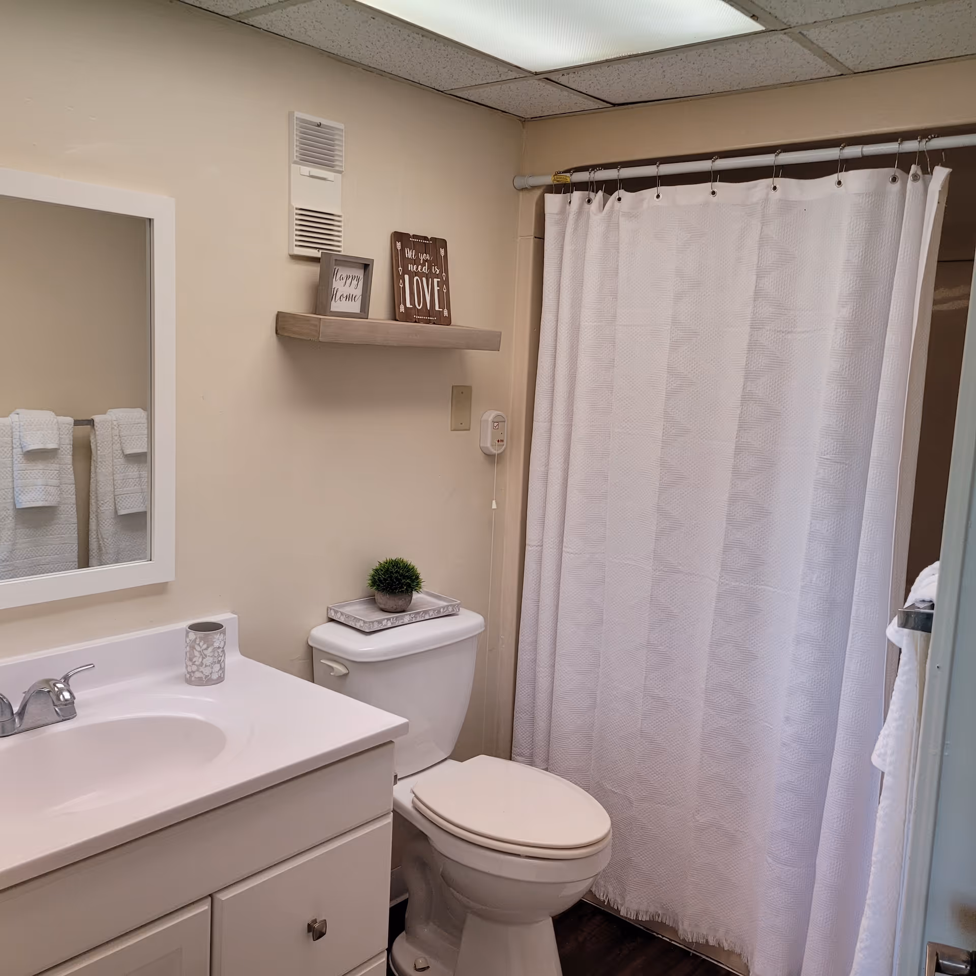 A clean bathroom with a white sink and cabinet, a toilet with a small plant and tray on top, a white shower curtain, a mirror above the sink, two white towels hanging on a rack, and a small wooden shelf on the wall holding two decorative signs.