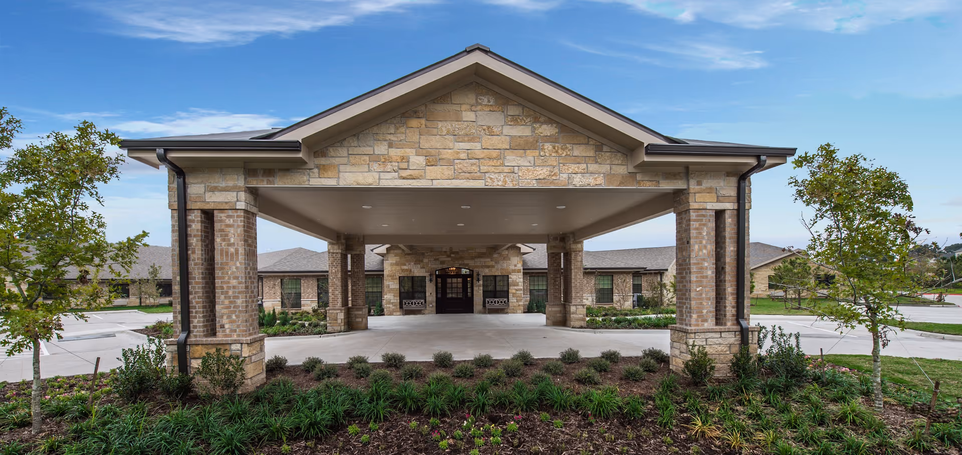 Covered main entrance and porte-cochère of a brick and stone senior living facility with landscaping and a driveway.