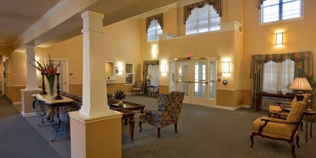 Spacious, warmly lit lobby/lounge with upholstered chairs, decorative columns, a table with flowers, and glass double entrance doors.