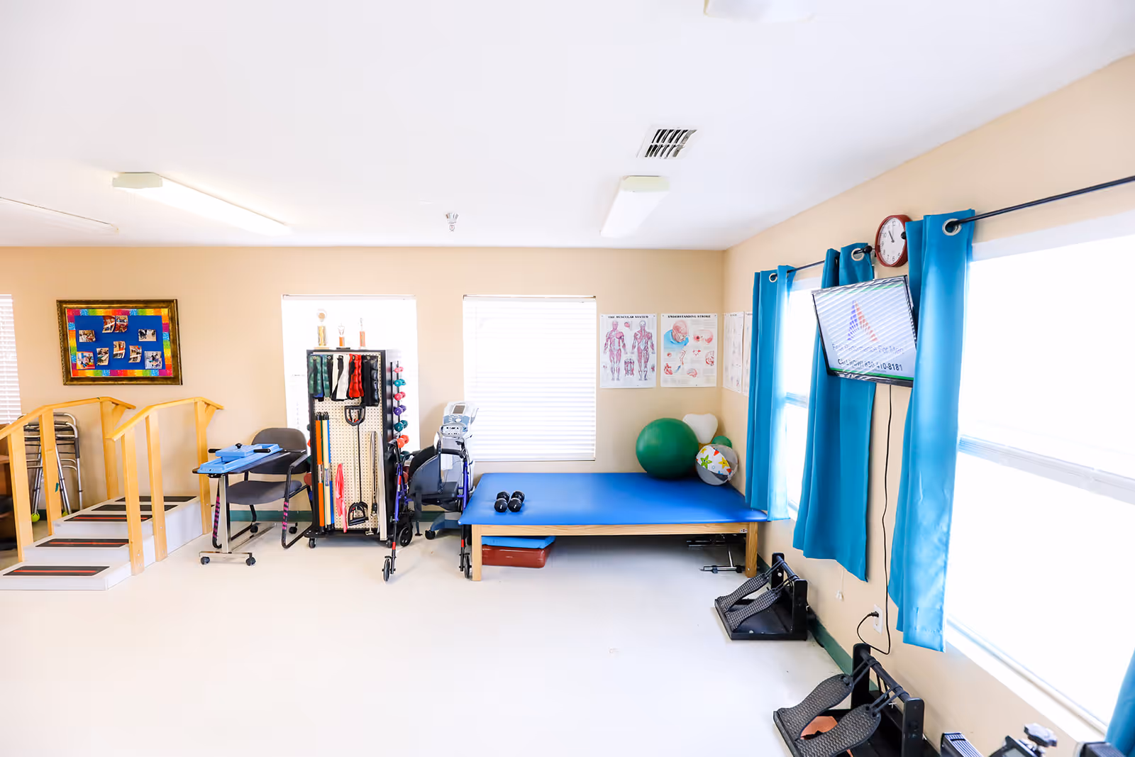 Bright physical therapy room with a treatment table, exercise equipment, practice stairs, exercise balls, anatomical posters and a wall-mounted TV.