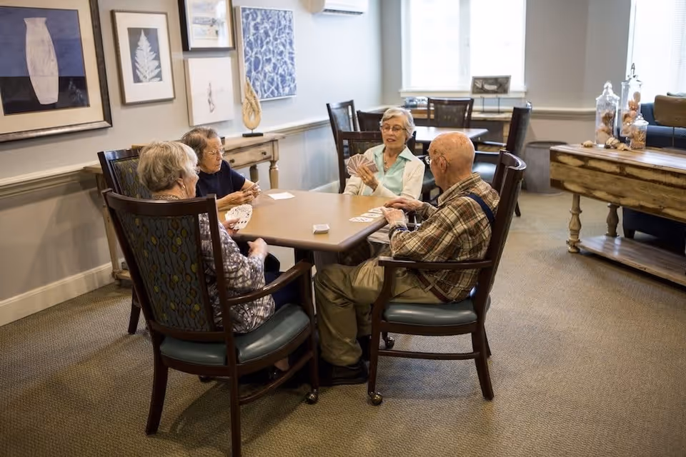 Four elderly people sitting around a square table playing cards in a well-lit room with framed artwork on the walls and wooden furniture.