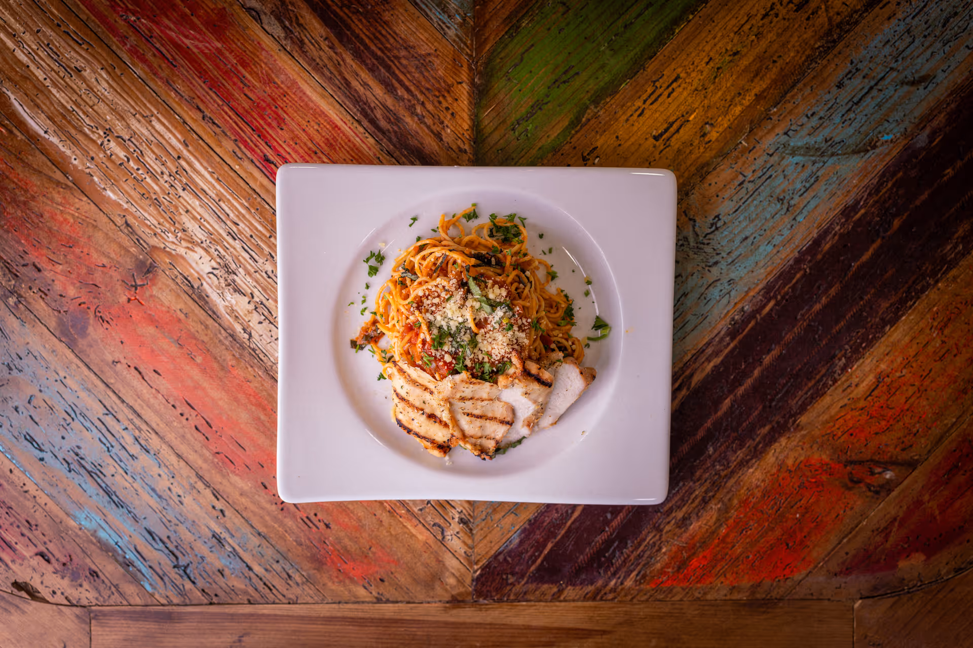 A square white plate with grilled chicken slices served on a bed of spaghetti pasta topped with tomato sauce, grated cheese, and chopped herbs, placed on a colorful wooden table with a chevron pattern.