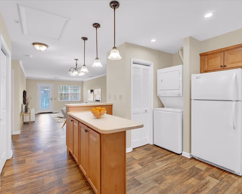 Interior view of a kitchen area with wooden cabinets, a white refrigerator, and a stacked washer and dryer unit. There is a kitchen island with a bowl of oranges on top, pendant lights hanging from the ceiling, and a view into a living area with a door and window in the background.