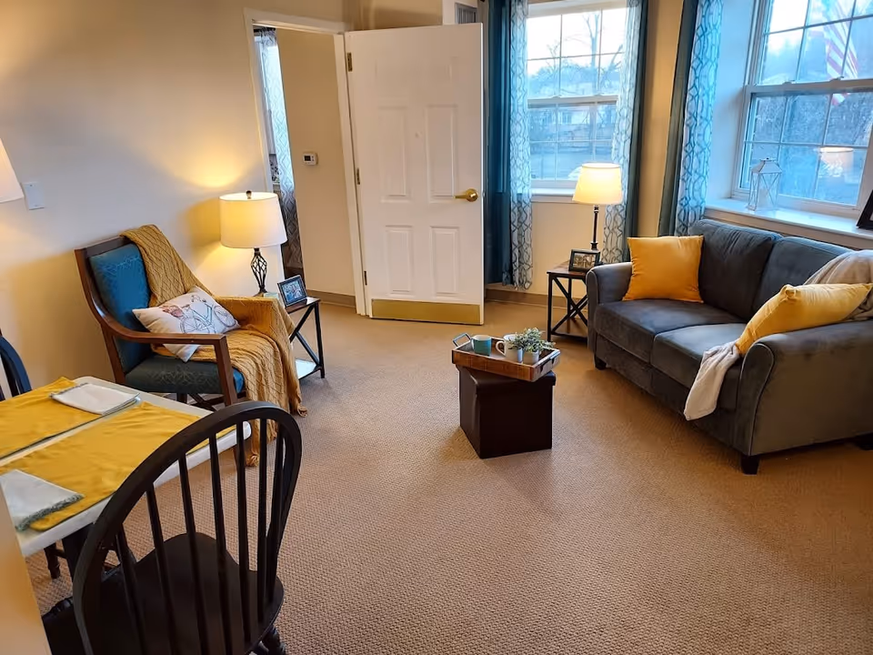 A cozy living room in a senior living facility with a gray sofa adorned with yellow pillows, a wooden armchair with a blue cushion and a yellow throw blanket, two side tables each with a lamp and framed photo, a small coffee table with a tray holding mugs and a small plant, and a dining table with yellow placemats and black chairs. The room has beige carpet, light-colored walls, and two windows with blue patterned curtains letting in natural light.