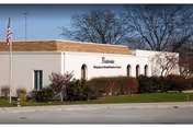 Exterior view of a single-story building with a flat roof and beige walls, surrounded by bushes and trees. An American flag is visible on a flagpole to the left. The building has a sign that reads 'Pembrooke Outpatient Rehabilitation Center.'