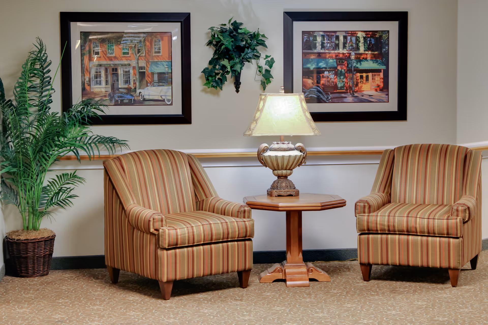 Two striped armchairs with a wooden side table and a decorative lamp between them, flanked by a potted plant on the left. Two framed paintings and a small wall-mounted plant are displayed on the beige wall behind the chairs.