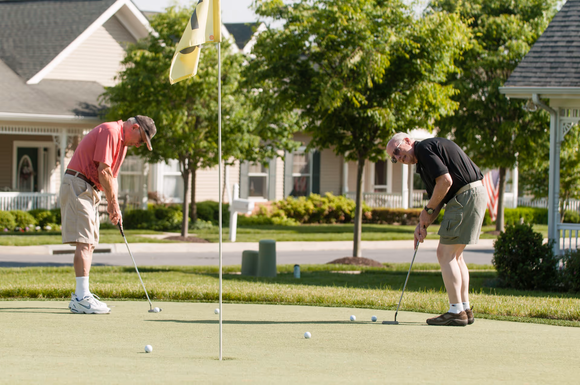 Two elderly men playing golf on a putting green in a residential community with houses and trees in the background.