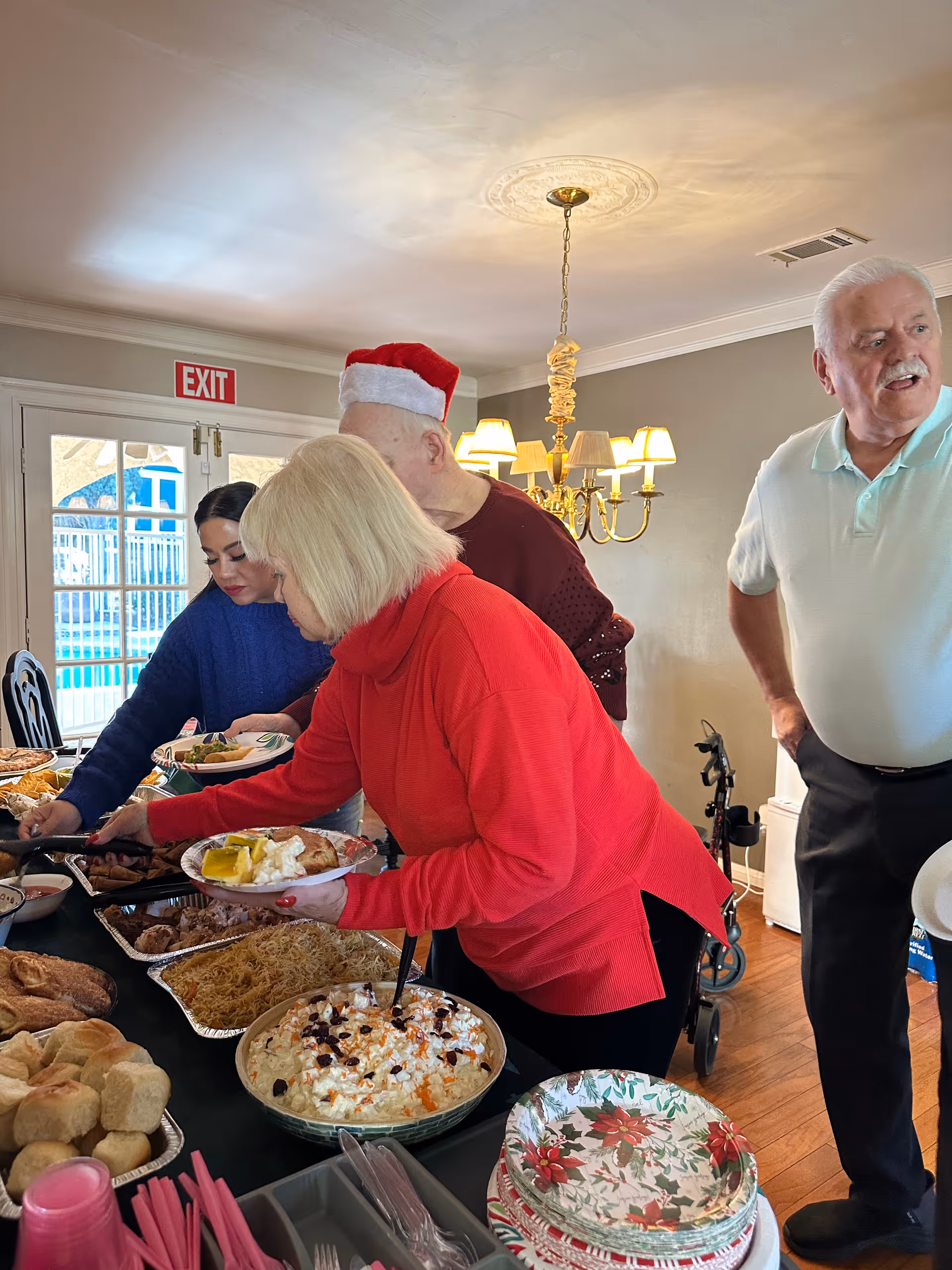 Several elderly people and a younger woman serving themselves food from a buffet table inside a room with a chandelier and an exit door in the background. The table is filled with various dishes including rolls, noodles, and a salad. One elderly man is wearing a Santa hat.