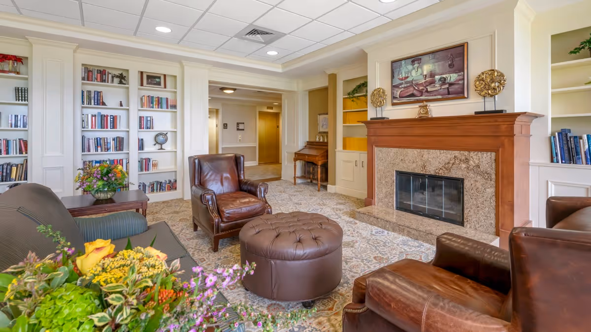 A comfortable common living room with leather chairs, a tufted ottoman, fireplace, built-in bookshelves and floral arrangements.