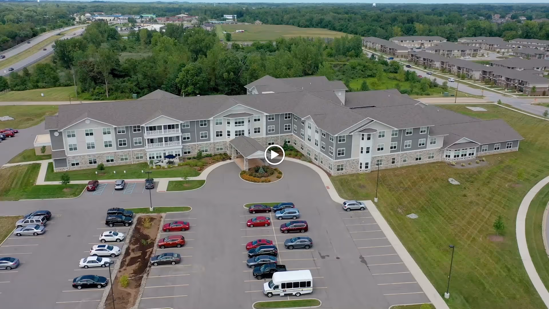 Aerial view of a large, three-story senior living facility named Residences at Coffee Creek, surrounded by green lawns and trees. The building has a gray roof and light-colored exterior walls with multiple windows. There is a parking lot in front with several cars and a white van parked. Roads and additional buildings are visible in the background.