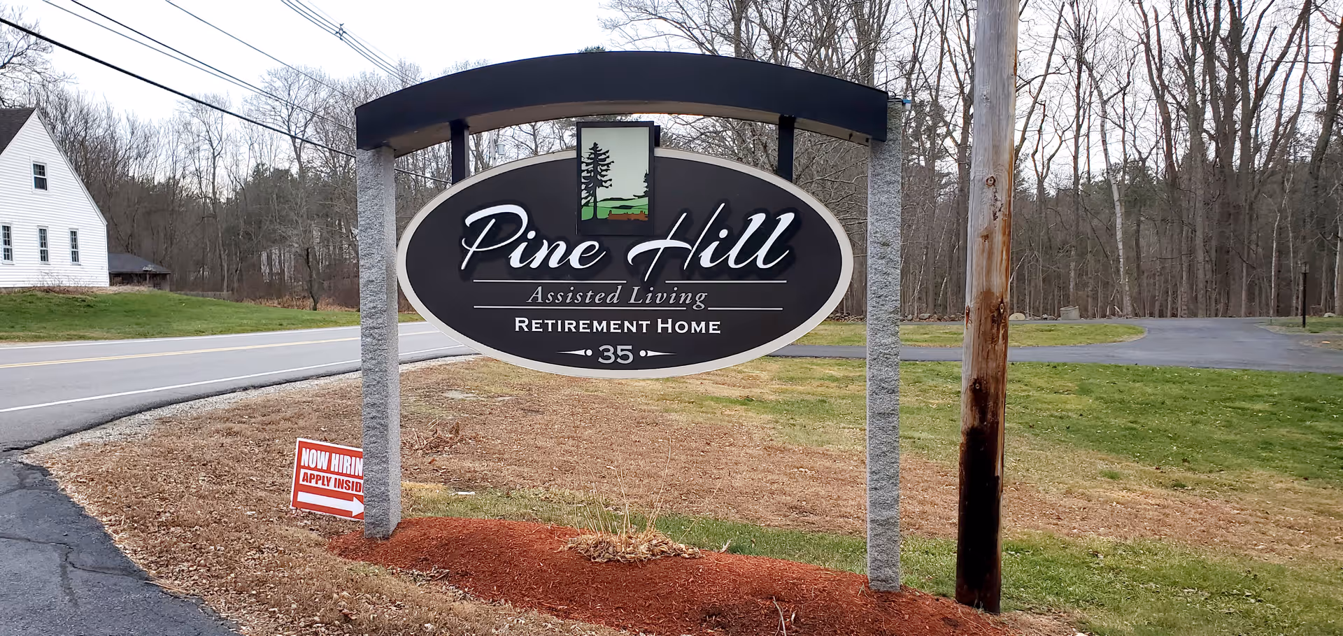 Outdoor view of a large black and white sign for Pine Hill Assisted Living Retirement Home, located near a road with trees and grass in the background. A smaller red and white sign nearby reads 'Now Hiring Apply Inside.'