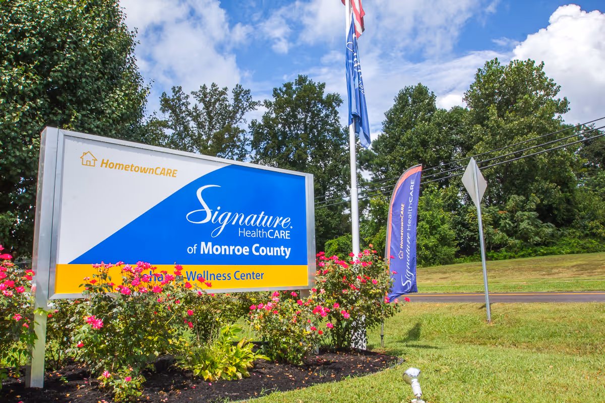Outdoor view of a sign for Signature HealthCARE of Monroe County Rehab & Wellness Center surrounded by pink flowers and greenery, with a flagpole and a blue flag nearby under a partly cloudy sky.