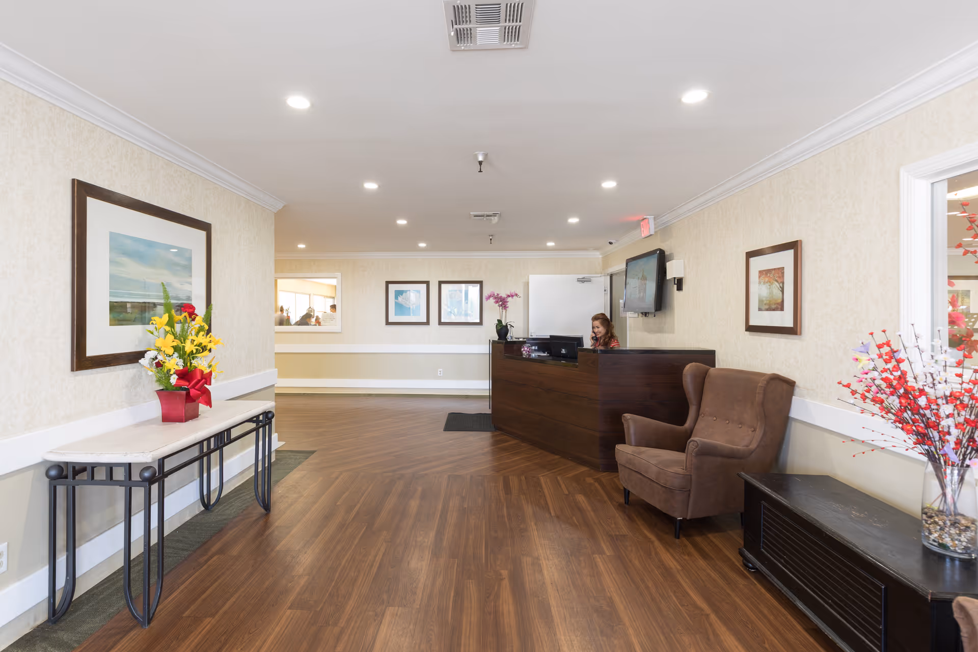 Reception area of a healthcare facility with a wooden reception desk where a woman is seated working on a computer. The room has wood flooring, beige walls with framed artwork, a brown upholstered armchair, and decorative flower arrangements on a console table and a side table.