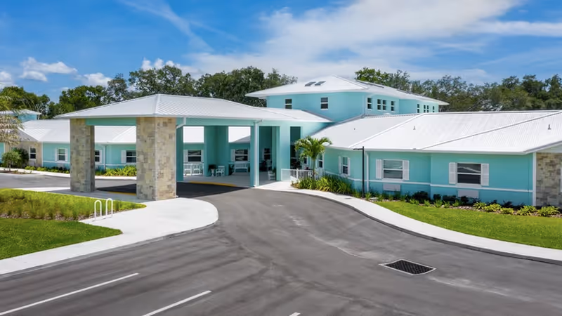 Front exterior of a light turquoise assisted living facility showing a covered entrance porte-cochere, surrounding grounds, and paved driveway.