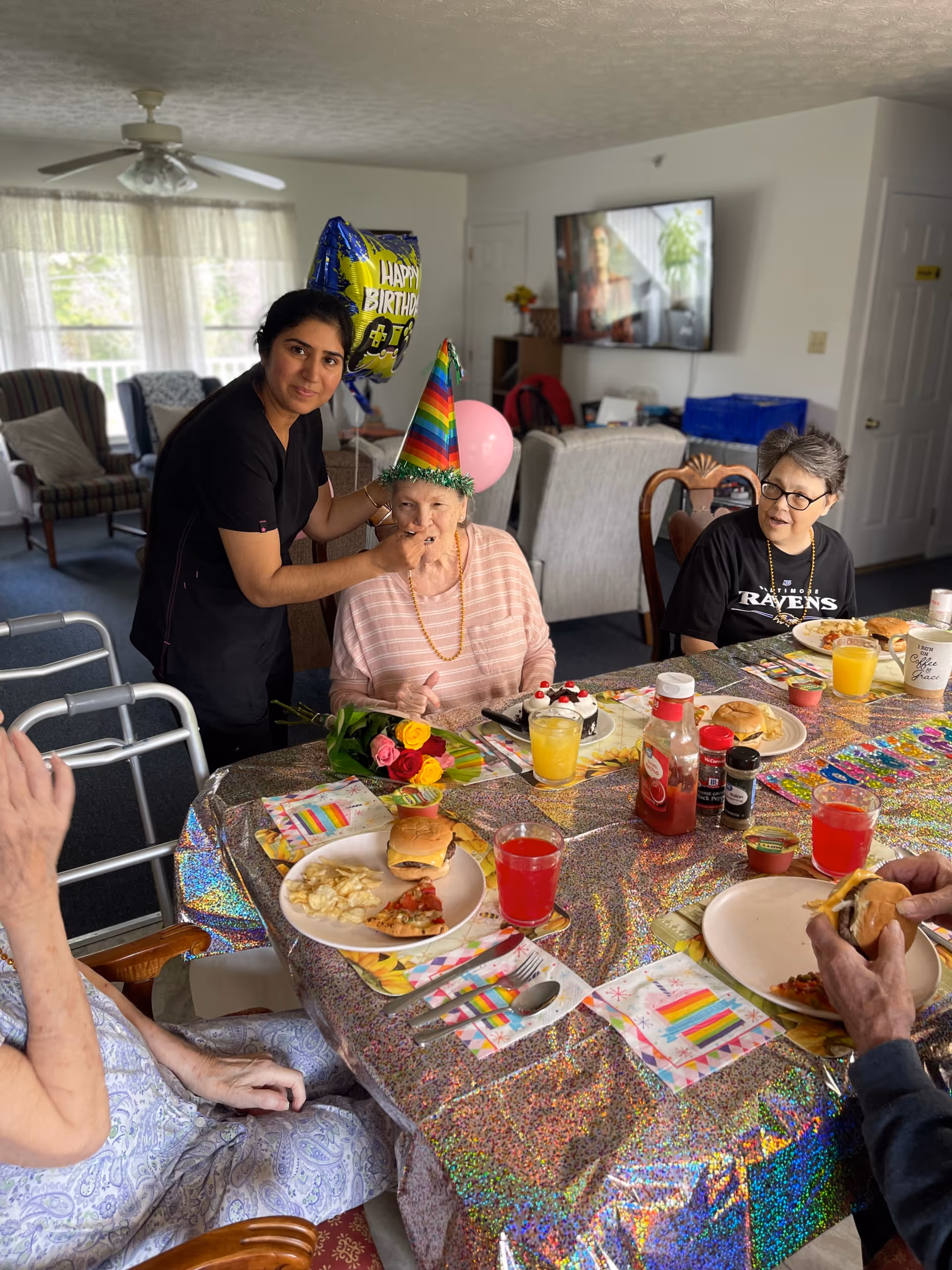 Residents and a caregiver celebrate a birthday around a table with party hats, balloons, cake and plates of food in a communal dining room.