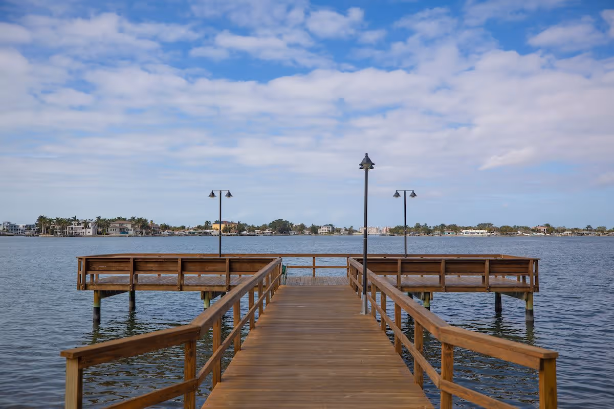 A wooden pier extending over a calm body of water with railings on both sides and three lamp posts along the pier. In the background, there are houses and trees along the shoreline under a partly cloudy sky.
