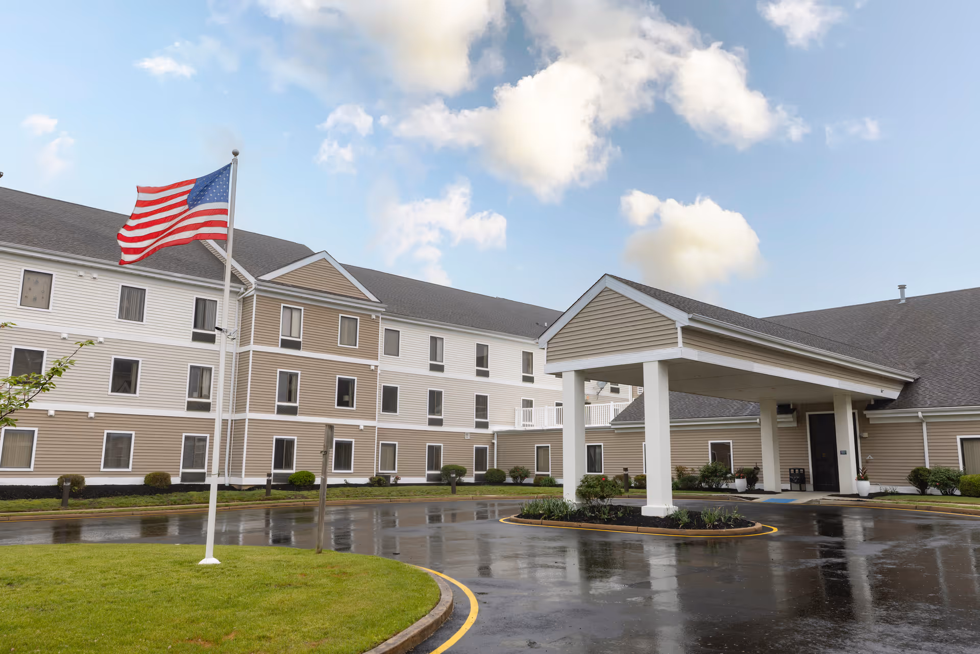 Exterior view of Mira Vie at Toms River assisted living facility showing a three-story building with beige and white siding, an American flag on a flagpole, a covered entrance, and a wet driveway under a partly cloudy sky.
