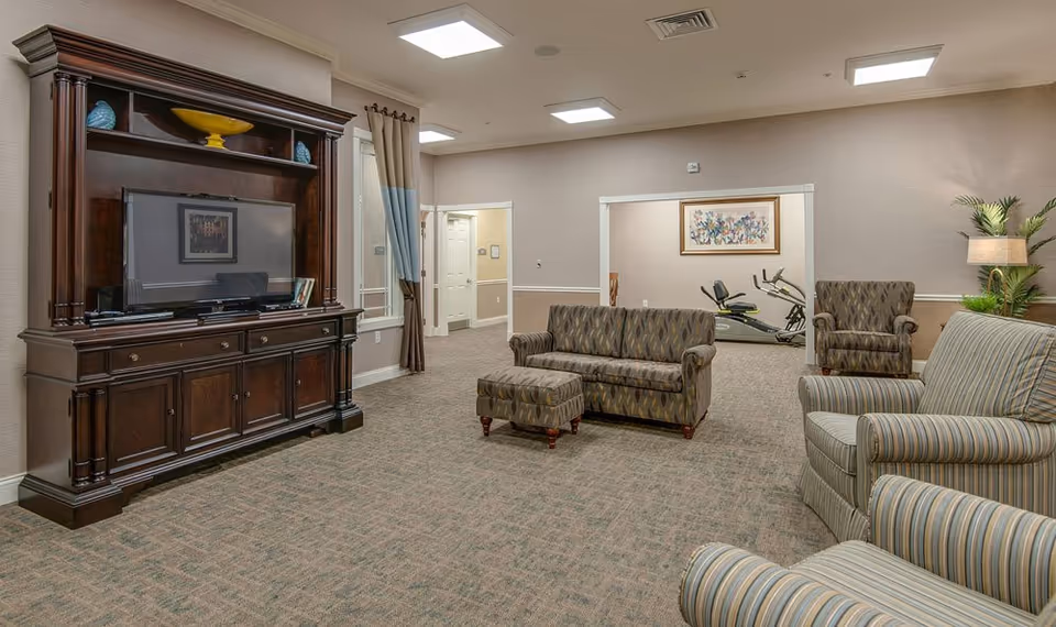 A spacious senior living facility common area with patterned upholstered sofas and armchairs arranged around a large wooden entertainment center with a flat-screen TV. The room has beige walls, carpeted floors, and recessed ceiling lights. In the background, there is an exercise bike visible through an open doorway, along with a framed artwork on the wall and a potted plant with a floor lamp.