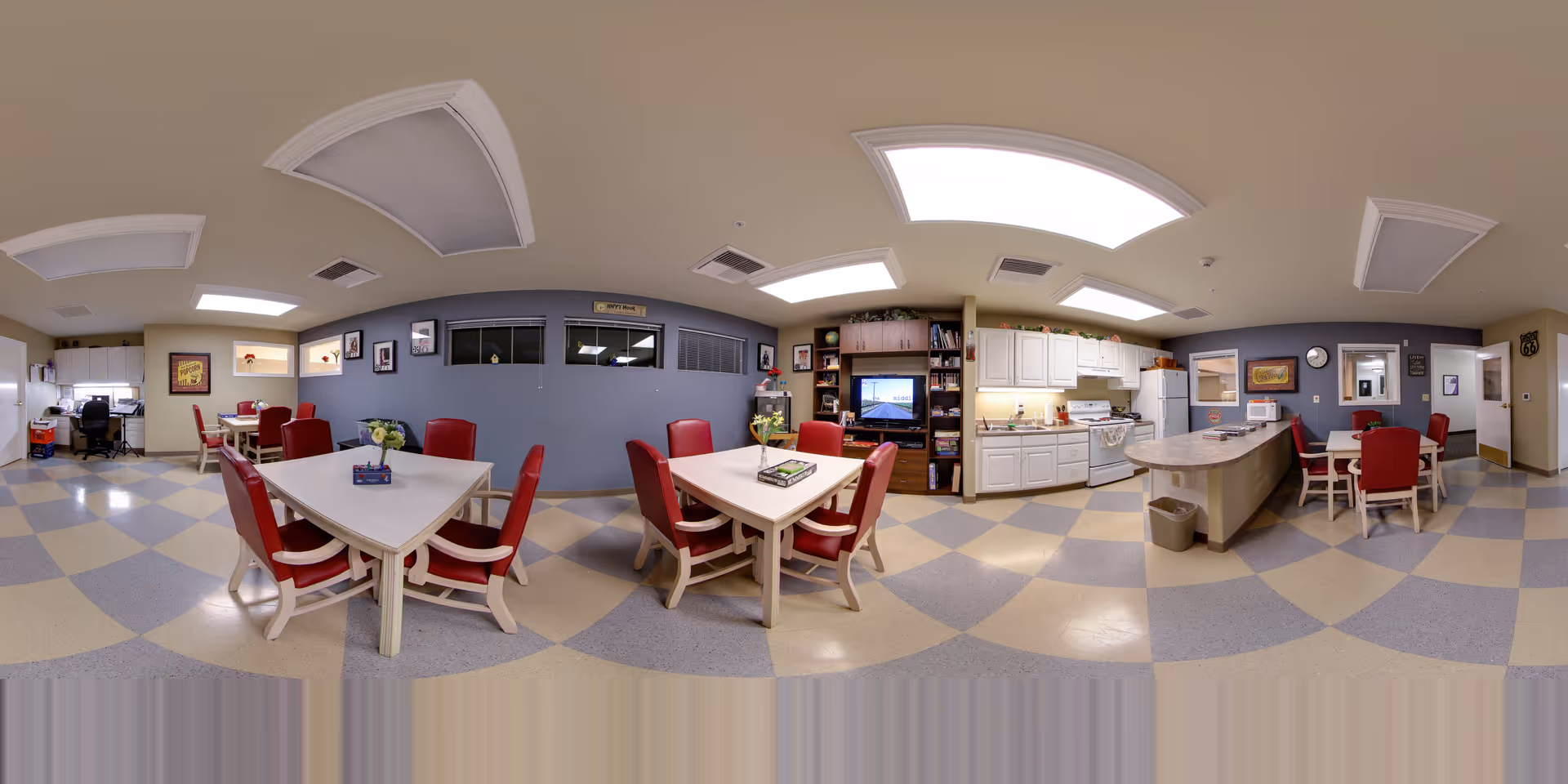 Communal dining and activity room with several tables and red chairs, a kitchenette, and a television.