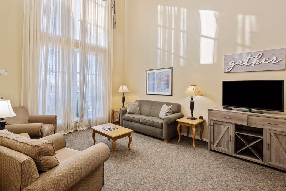 A cozy living room with a gray sofa, two beige armchairs, a wooden coffee table, two side tables with lamps, a wooden TV stand with a flat-screen TV, and a wall decoration that says 'gather'. Large windows with sheer curtains allow natural light to fill the room.
