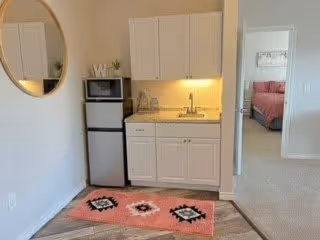 Small kitchenette area with white cabinets, a microwave, and a mini refrigerator. There is a round mirror on the wall to the left and a pink patterned rug on the floor. Through an open doorway, a bedroom with a bed and pink bedding is visible.