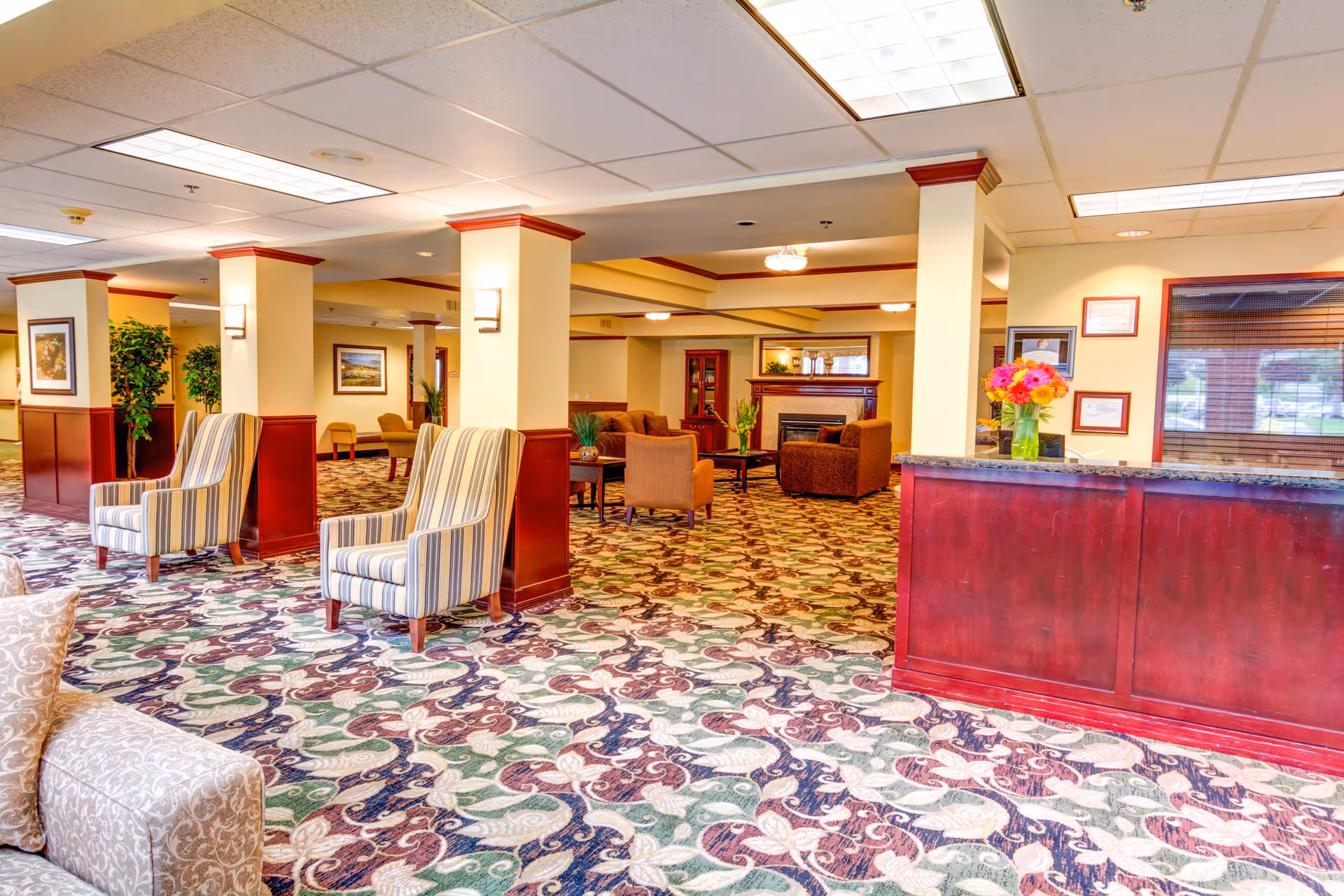 A spacious and well-lit senior living facility common area with patterned carpet flooring, several upholstered armchairs, a wooden reception desk with a vase of colorful flowers, framed artwork on the walls, and a seating area with sofas and a fireplace in the background.