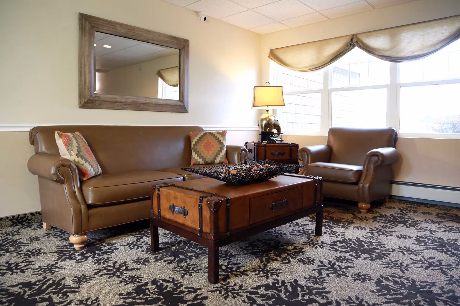 A cozy living room area with a brown leather sofa and matching armchair, both adorned with patterned throw pillows. A wooden coffee table with metal accents sits in front of the sofa, topped with a decorative black wire bowl filled with ornamental balls. A table lamp with a beige shade and an elephant-shaped base is placed on a small side table near the window. The room features a large window with beige curtains, a patterned carpet, and a large wooden-framed mirror on the wall.