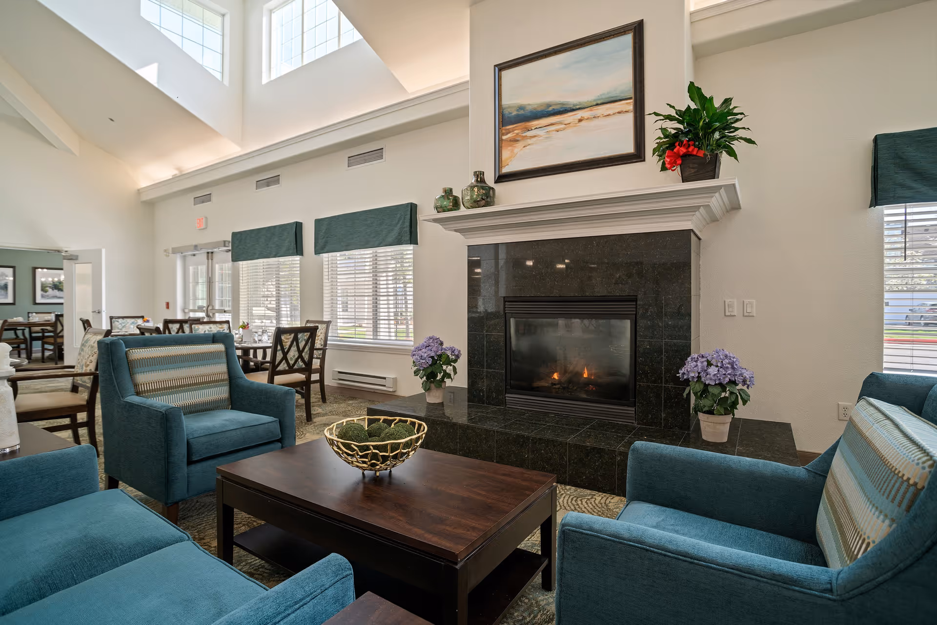 A cozy living room area in a senior living facility with teal upholstered armchairs and a matching sofa arranged around a dark wooden coffee table. The room features a black tiled fireplace with a fire burning, decorated with potted plants and a landscape painting above the mantel. Large windows with green valances let in natural light, and a dining area with tables and chairs is visible in the background.